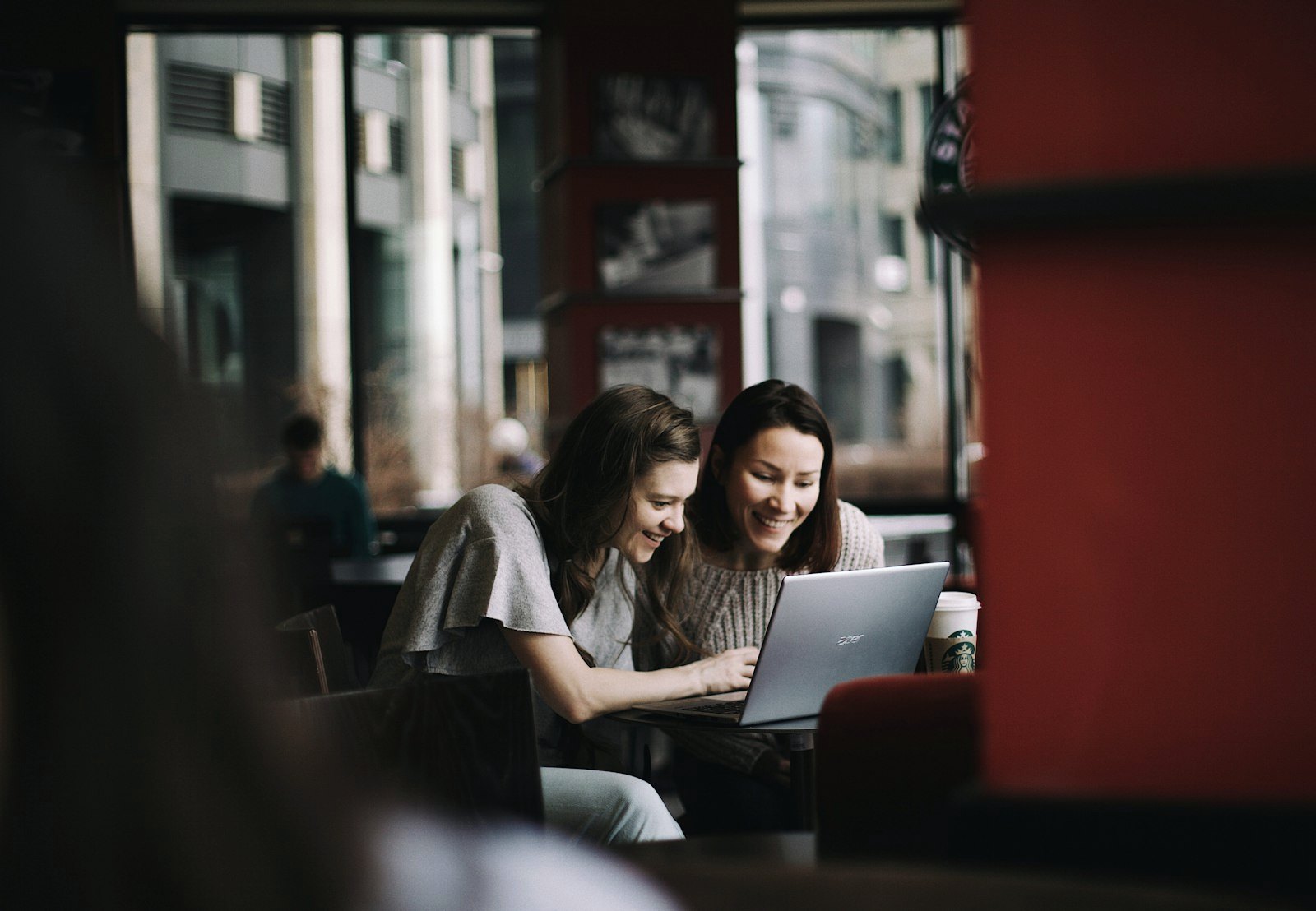 Duas mulheres sorrindo diante de um notebook numa mesa perto da janela.
